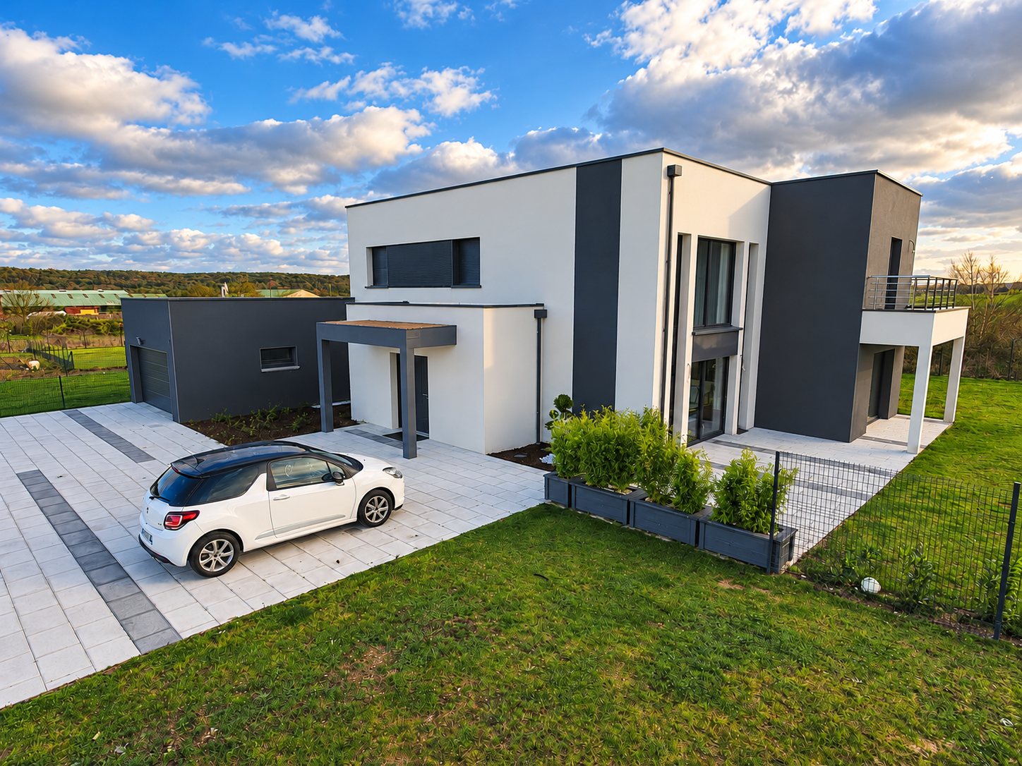 Une maison contemporaine blanche et grise à toit plat, avec garage, terrasse et allée pavée où est garée une voiture blanche.