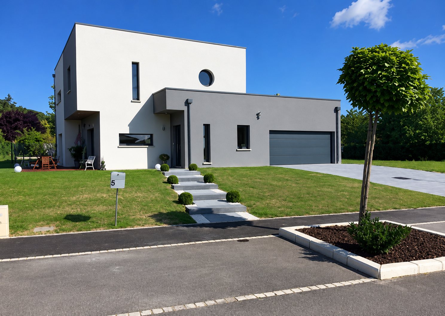 Maison moderne de style cubique aux façades blanche et grise, avec garage gris foncé, pelouse verte et escalier en pierre sous un ciel bleu.