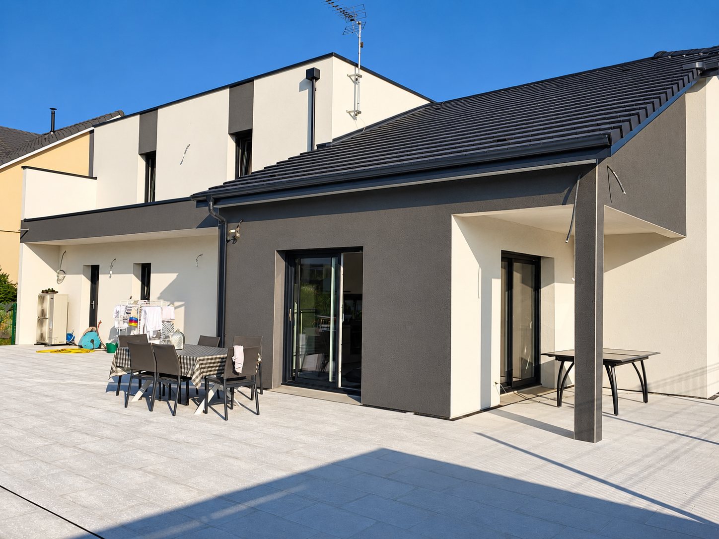 Façade d'une maison moderne aux enduits gris anthracite et blanc cassé, toit en tuiles noires et grande terrasse carrelée avec table et chaises.