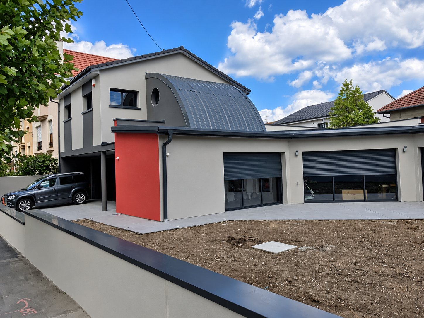 Façade d'une maison moderne aux murs gris, avec un toit en arc métallique, un pan rouge et deux garages à volets roulants, devant un terrain en terre.