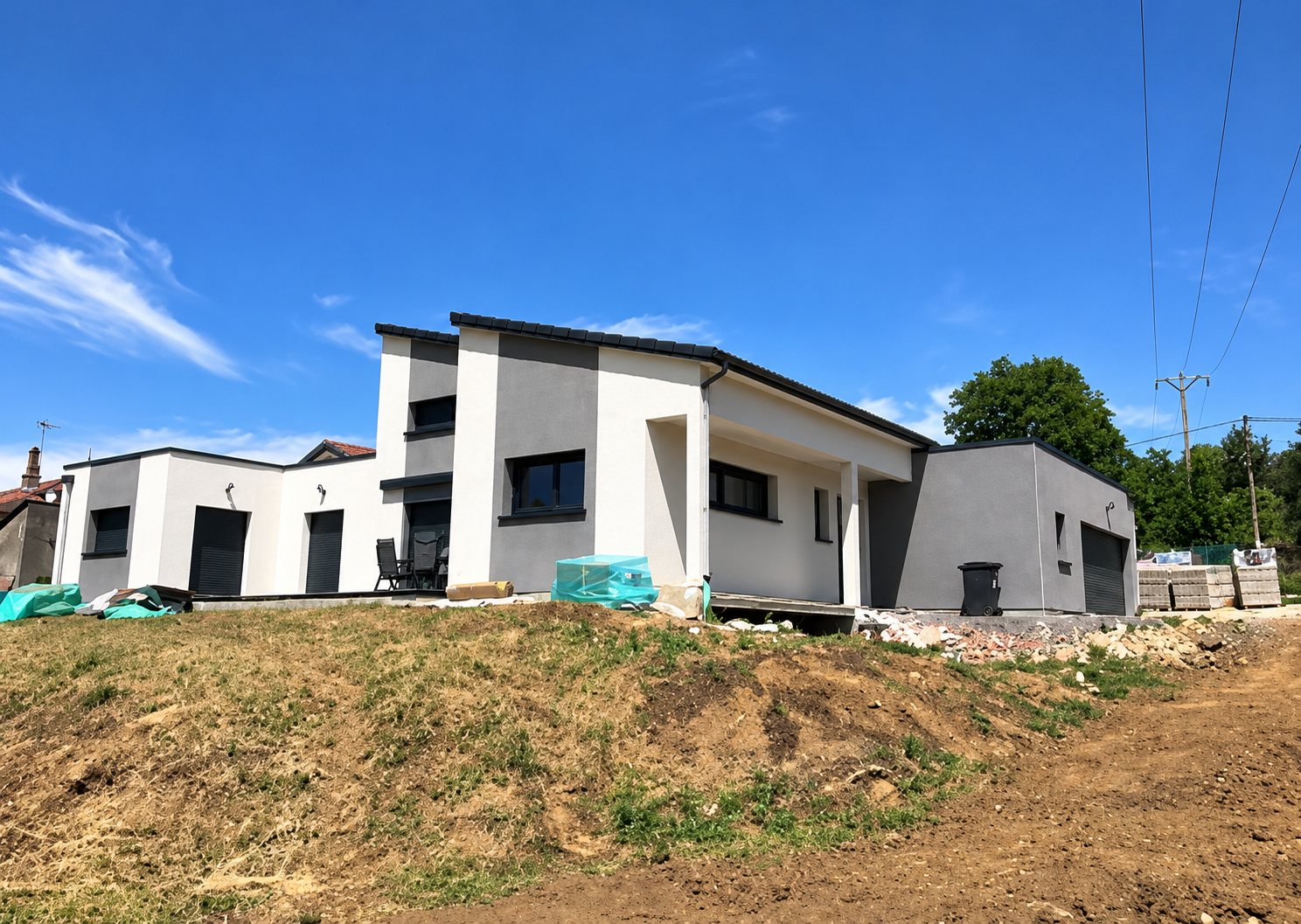 Maison moderne de plain-pied aux façades blanches et grises, en cours de finition, entourée de terre fraîchement remuée sous un ciel bleu.