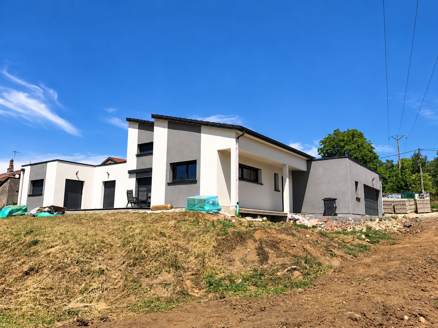 Maison moderne de plain-pied aux murs blancs et gris et aux menuiseries anthracite, photographiée sur un terrain de chantier sous un ciel bleu.