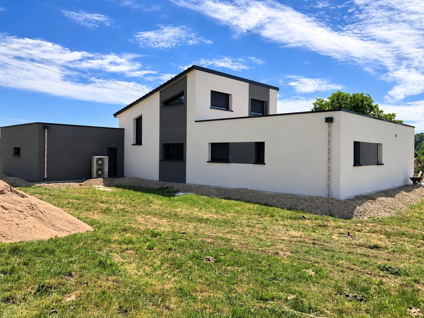 Maison moderne récemment construite aux murs enduits blancs et gris anthracite, avec pompe à chaleur extérieure et terrain en herbe au premier plan.