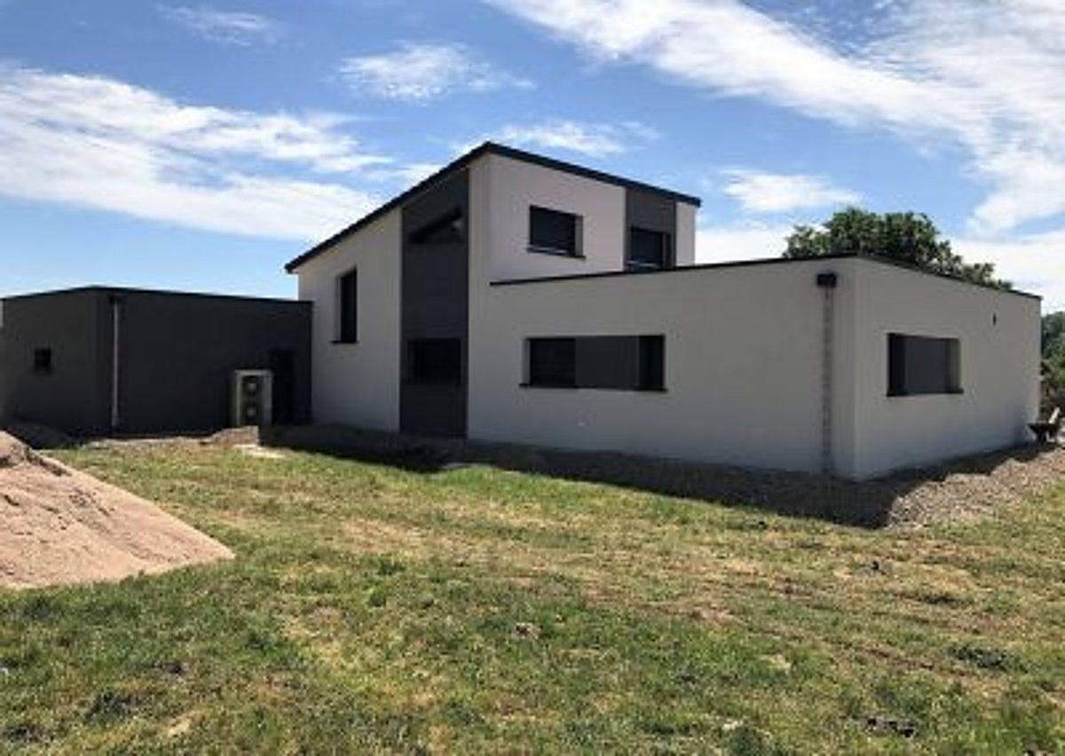 Maison moderne à toit plat, aux façades blanches et grises, entourée d'une pelouse sous un ciel bleu légèrement nuageux.