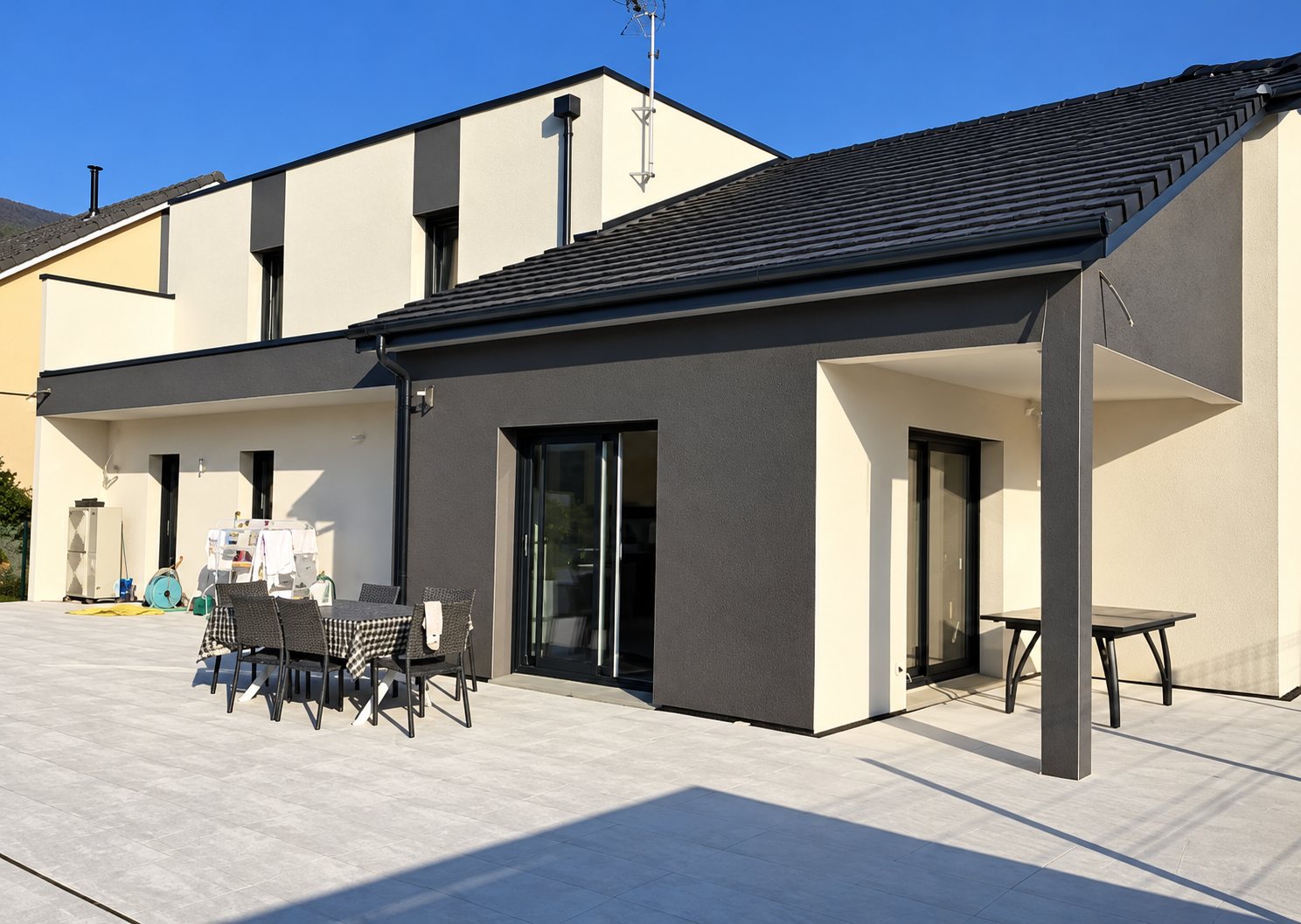 Une maison moderne aux façades grises et blanches, au toit sombre, avec une vaste terrasse carrelée où se trouvent une table et des chaises.
