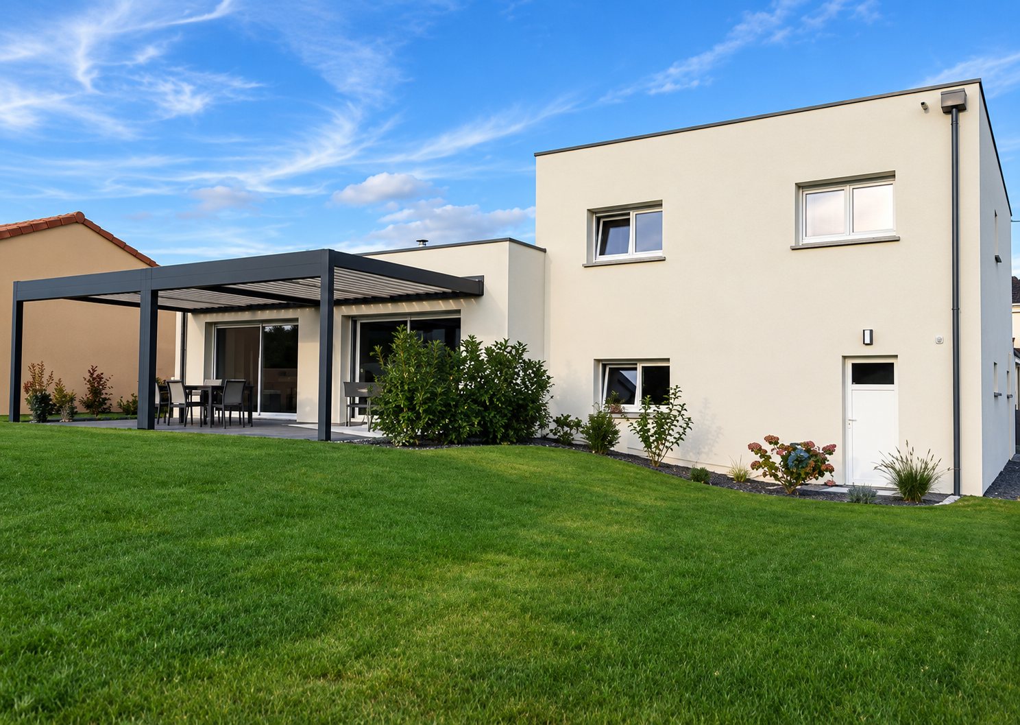 Maison moderne de couleur beige à toit plat, dotée d'une pergola sur une terrasse et d'un grand jardin engazonné sous un ciel bleu.
