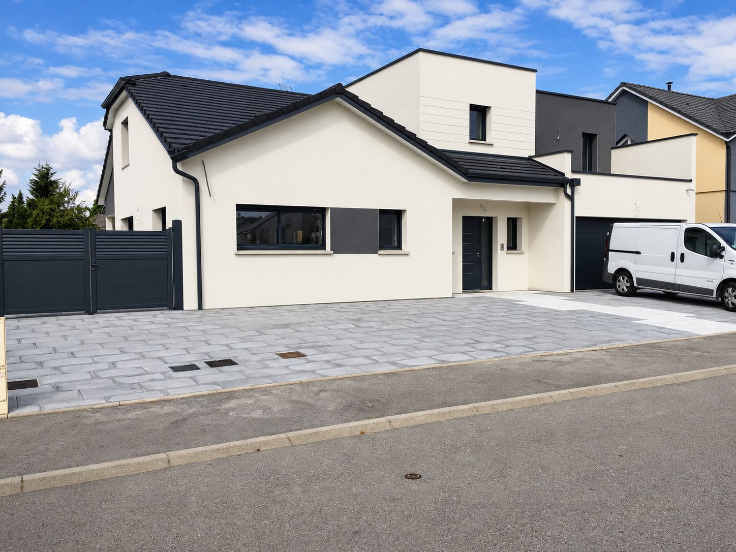 Maison contemporaine aux murs blancs et toit anthracite, dont l'allée est pavée de dalles grises, avec un portail gris et une camionnette blanche garée à droite.