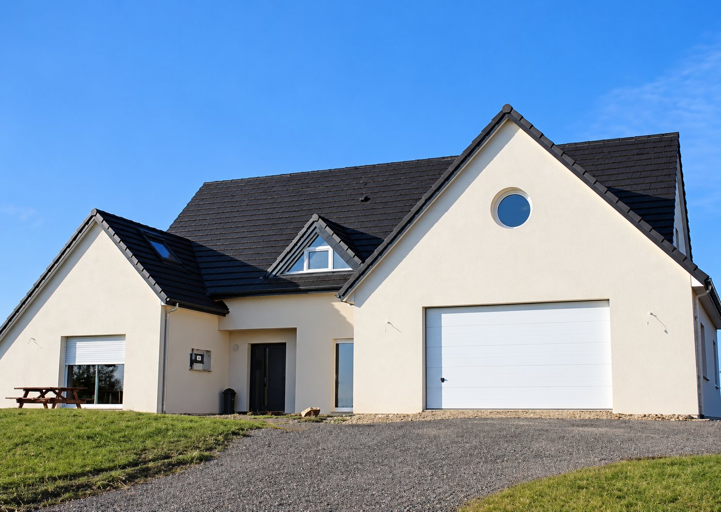 Une maison contemporaine aux murs blancs et au toit en ardoise sombre, avec une porte de garage blanche et une fenêtre ronde, sous un ciel bleu.
