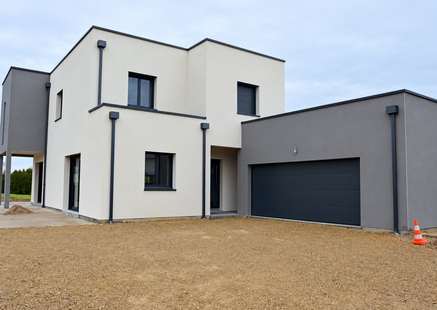 Maison contemporaine à toit plat aux façades blanches et grises, dotée d'un garage gris foncé et d'une allée en gravier au premier plan.