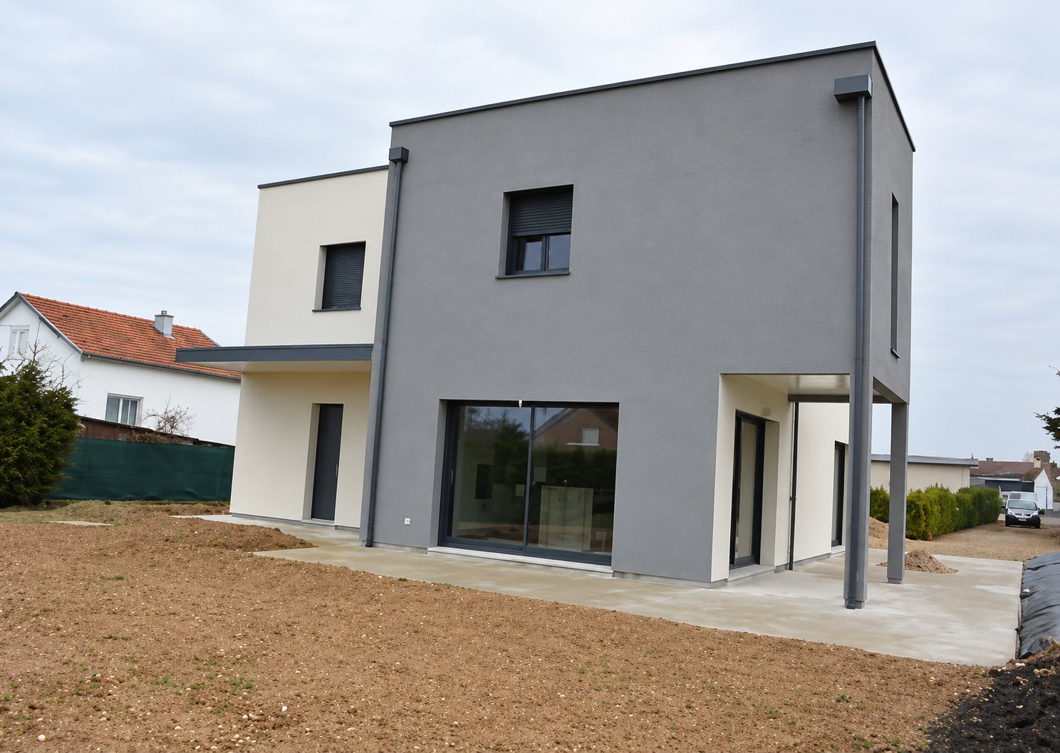 Maison individuelle contemporaine de forme cubique aux façades grises et blanches, entourée d'un terrain de terre non aménagé, sous un ciel couvert.