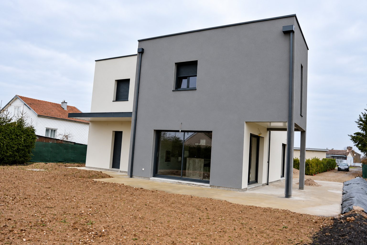 Maison contemporaine à toit plat aux façades grises et blanches, avec grande baie vitrée et terrasse en béton, entourée d'un terrain de terre nue.