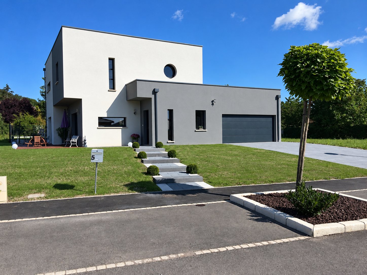 Maison contemporaine à toit plat, aux façades blanche et grise, avec garage anthracite, pelouse, boîte aux lettres numérotée 5 et escaliers en pierre grise menant à l'entrée.