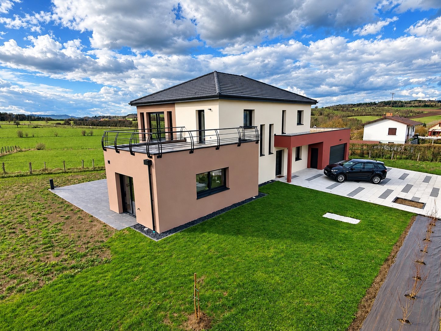 Vue aérienne d'une maison contemporaine à toit gris foncé, avec terrasse à garde-corps métallique, garage bordeaux et voiture noire, entourée de prairies.