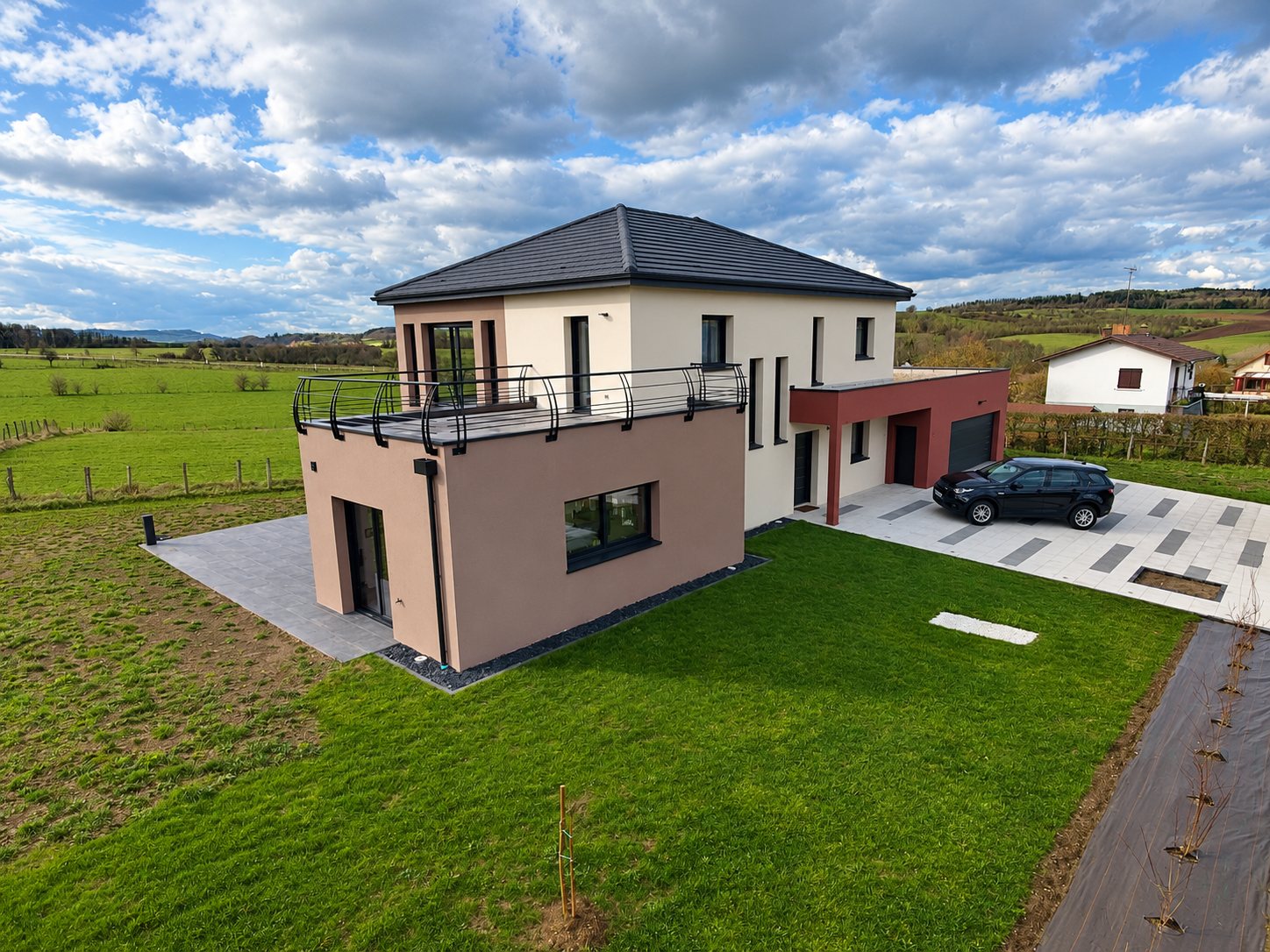 Vue aérienne d'une maison contemporaine à toit gris foncé, avec terrasse à garde-corps métallique, garage bordeaux et voiture noire, entourée de prairies.