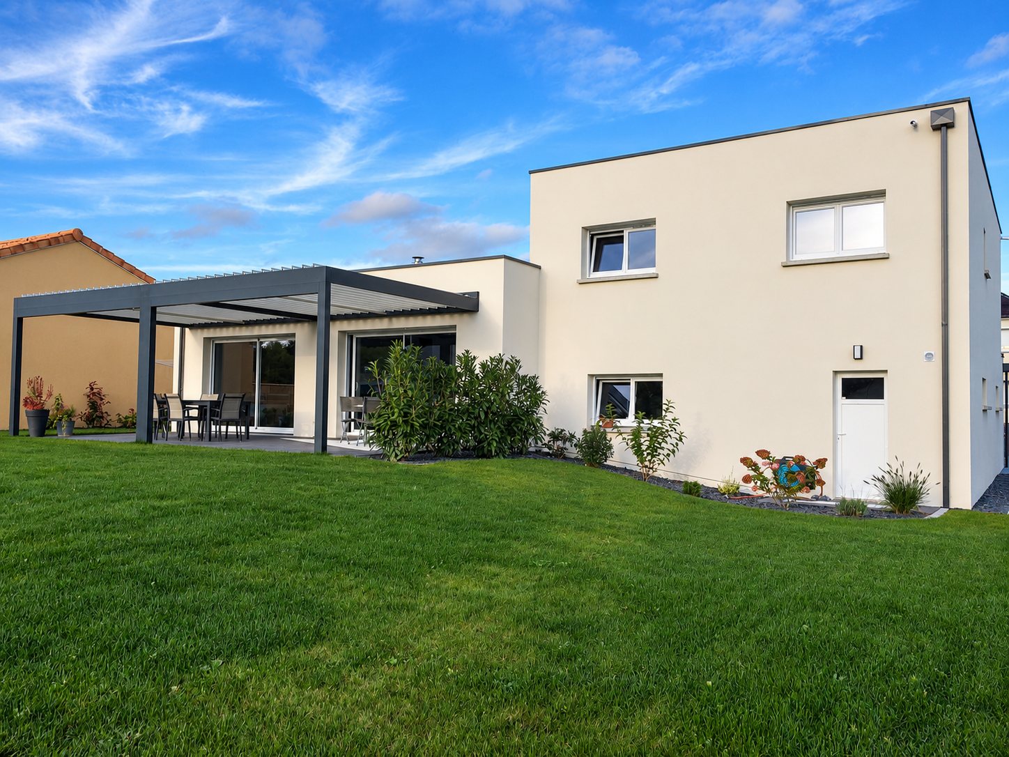 Maison contemporaine à toit plat de couleur crème avec une pergola bioclimatique grise couvrant une terrasse et une grande pelouse au premier plan.