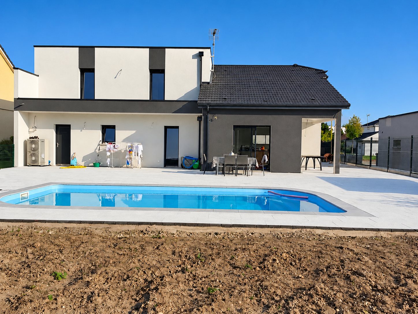 Façade arrière d'une maison contemporaine avec une piscine bleue entourée d'une grande terrasse blanche et d'un terrain de terre au premier plan.