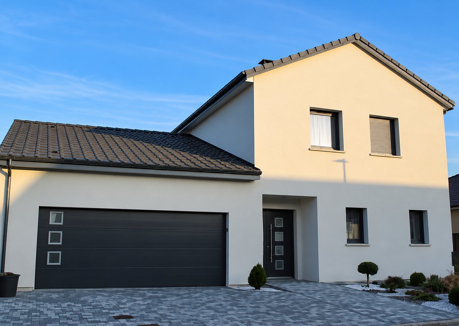 Façade d'une maison récente aux murs clairs et au toit sombre, équipée d'une grande porte de garage anthracite à hublots, devant une allée pavée.