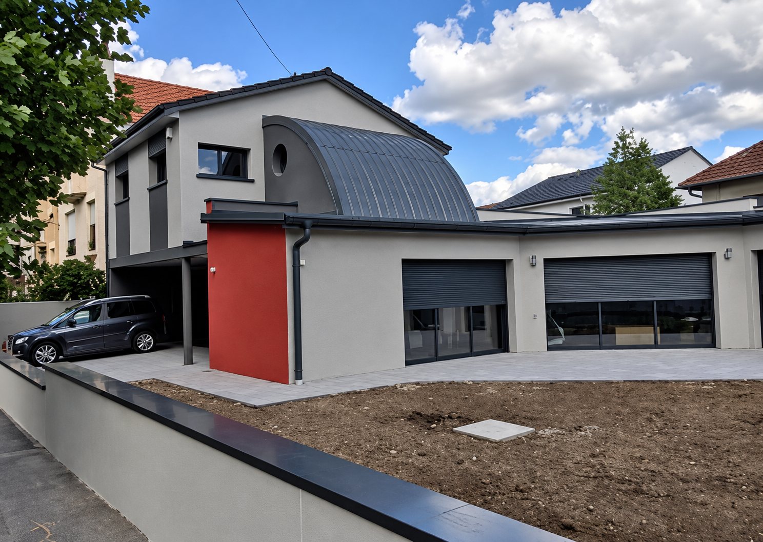 Maison contemporaine aux murs gris clair, avec un toit cintré en zinc, un panneau rouge et deux garages, devant un terrain fraîchement terrassé.