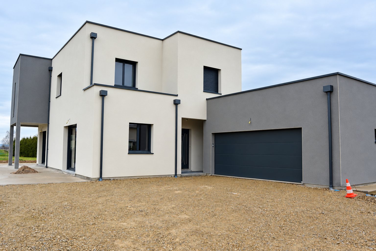 Maison contemporaine à toit plat, aux façades blanche et grise, avec un garage à porte sombre et une allée en gravier au premier plan.