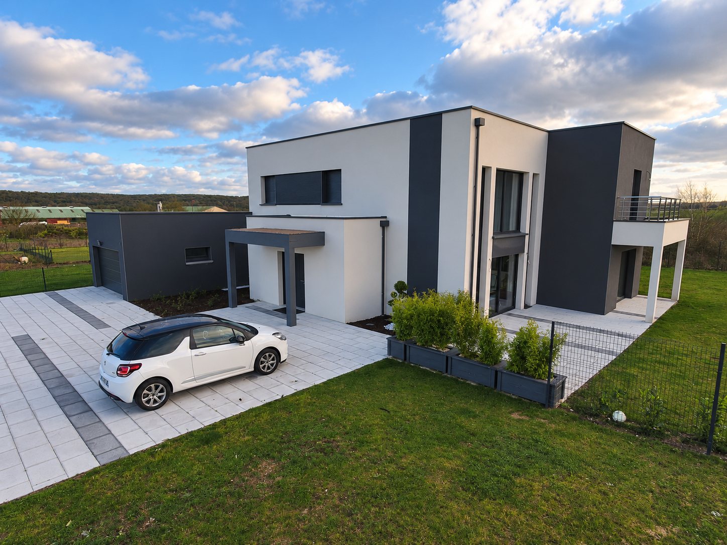 Une maison contemporaine blanche et grise à toit plat, avec garage, terrasse et allée pavée où est garée une voiture blanche.