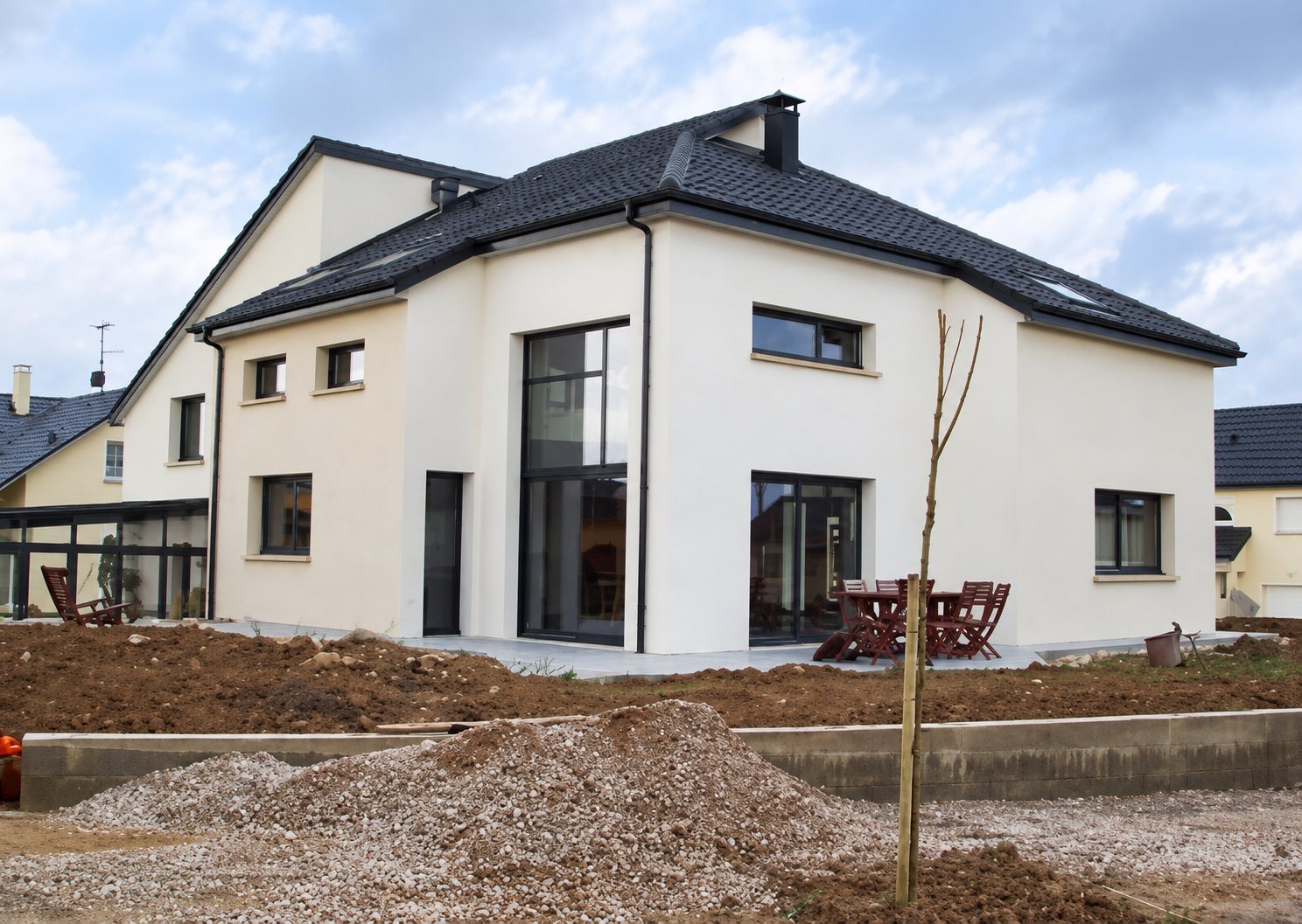 Maison individuelle récente aux façades blanches et au toit sombre, entourée de terre fraîchement remuée et d'un tas de gravats au premier plan.