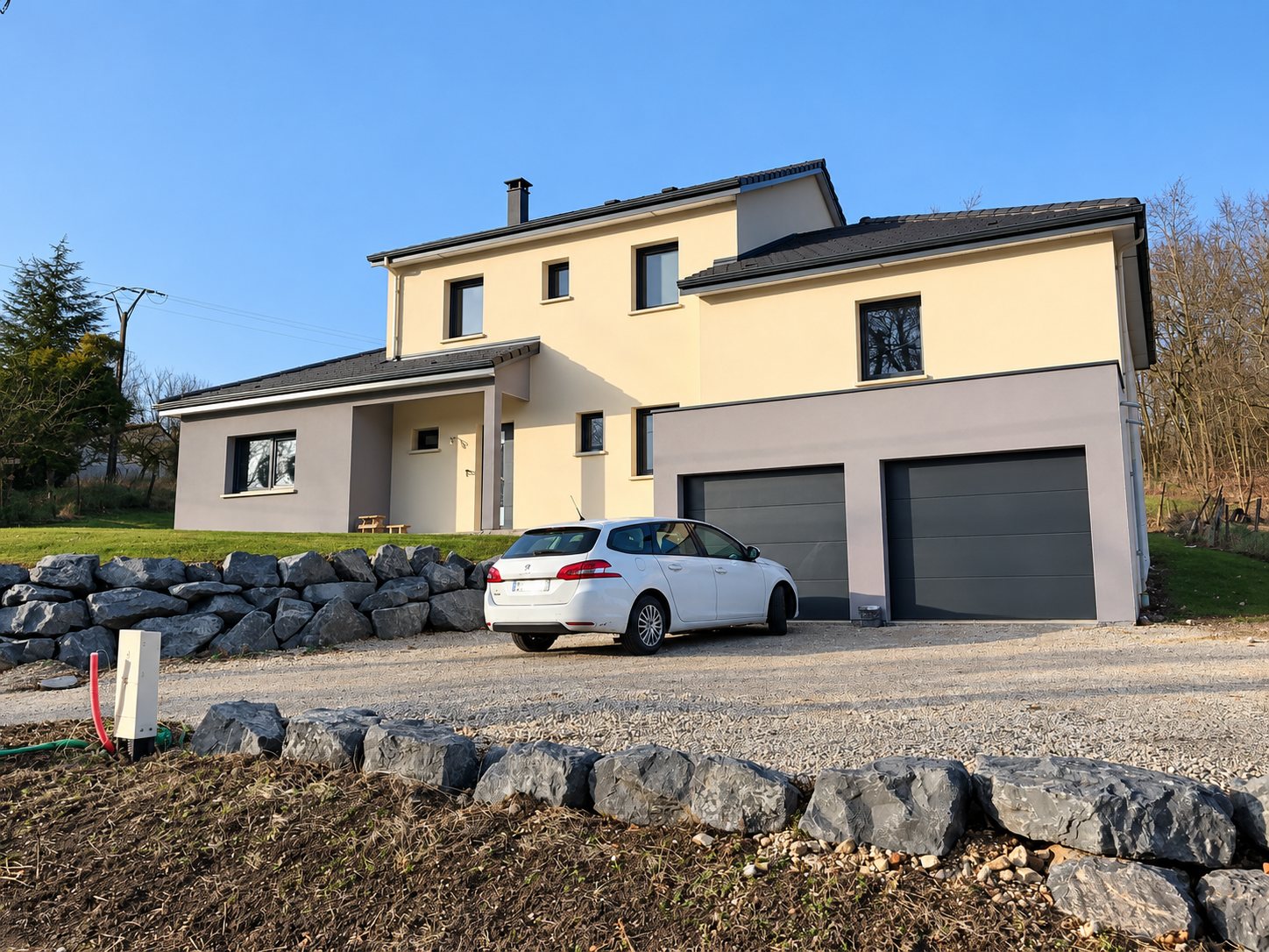 Maison récente à étage aux façades beige et grise, avec deux portes de garage grises, une voiture blanche sur l'allée de gravier et des murets de gros blocs de pierre.