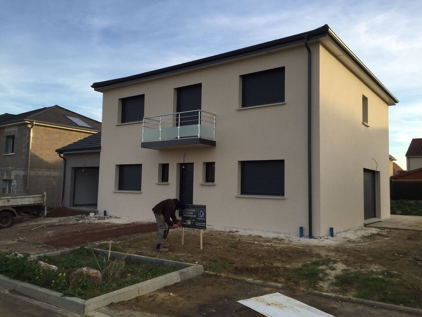 Une maison individuelle neuve à étage, à l'enduit beige et aux volets roulants gris, sur un terrain en chantier où un homme installe un panneau.