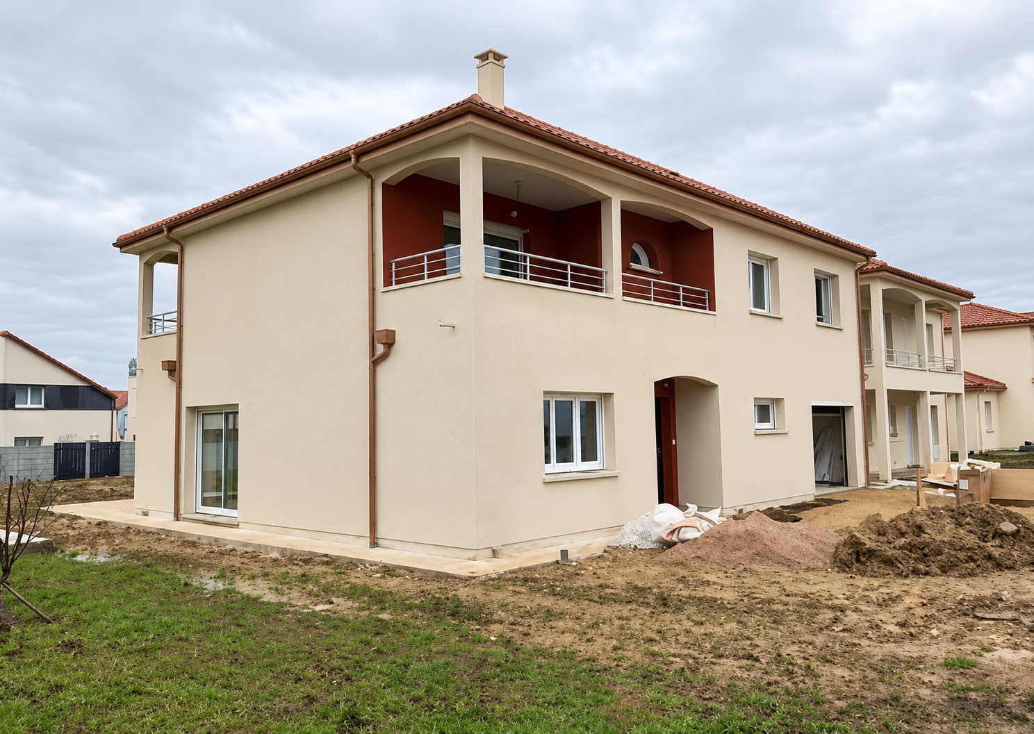 Une maison récente à étage aux murs beige clair et à la toiture rouge, entourée de terre fraîchement remuée sous un ciel couvert.