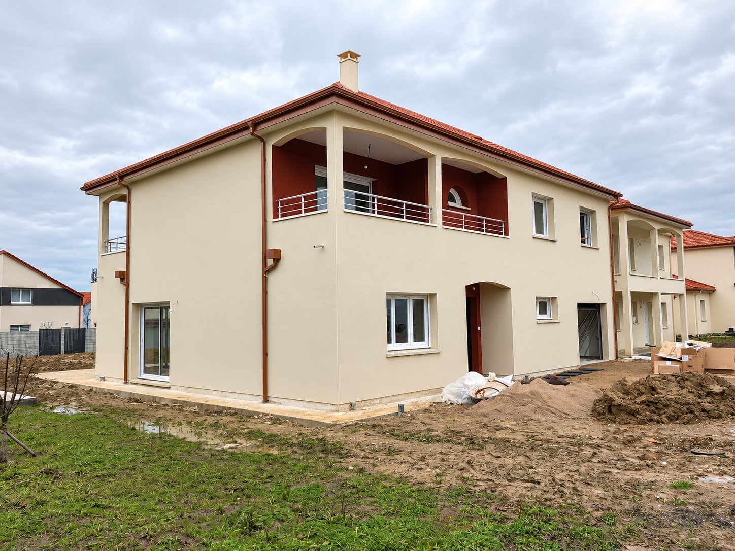 Maison neuve à étage avec façade crépie beige, balcon aux murs rouges et toit en tuiles, entourée d'un terrain boueux en cours de chantier.
