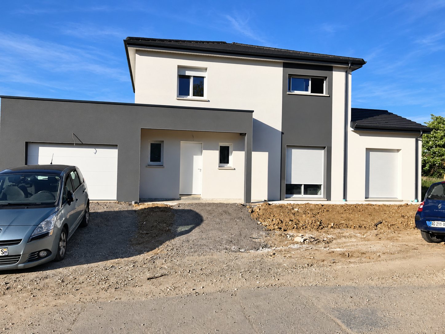 Maison récente à étage aux façades blanches et grises avec garage, volets roulants et toit sombre, terrain encore en terre devant.