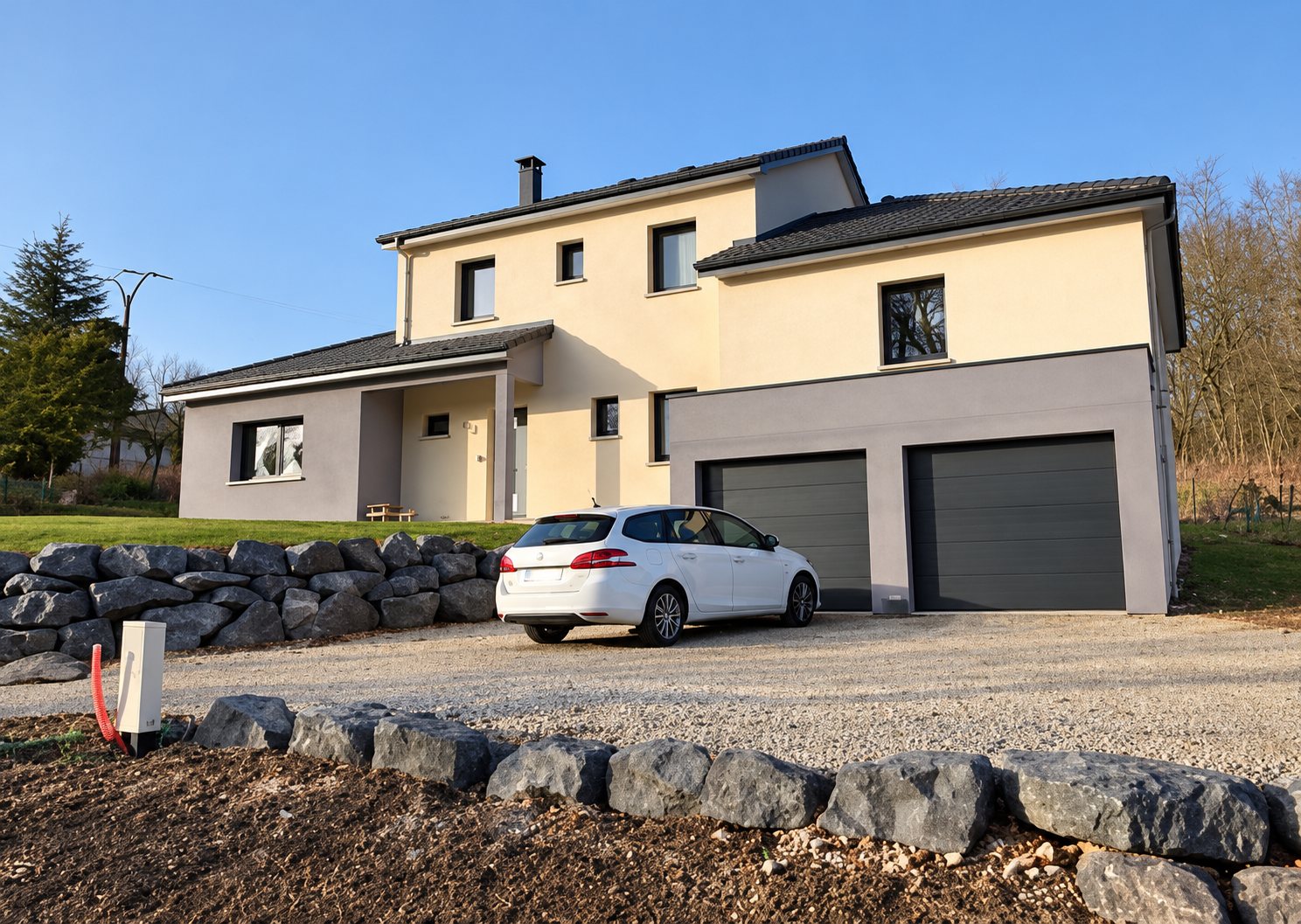 Maison récente à étage aux façades beige et grise, avec deux portes de garage, une voiture blanche garée sur le gravier et des gros blocs de pierre au premier plan.