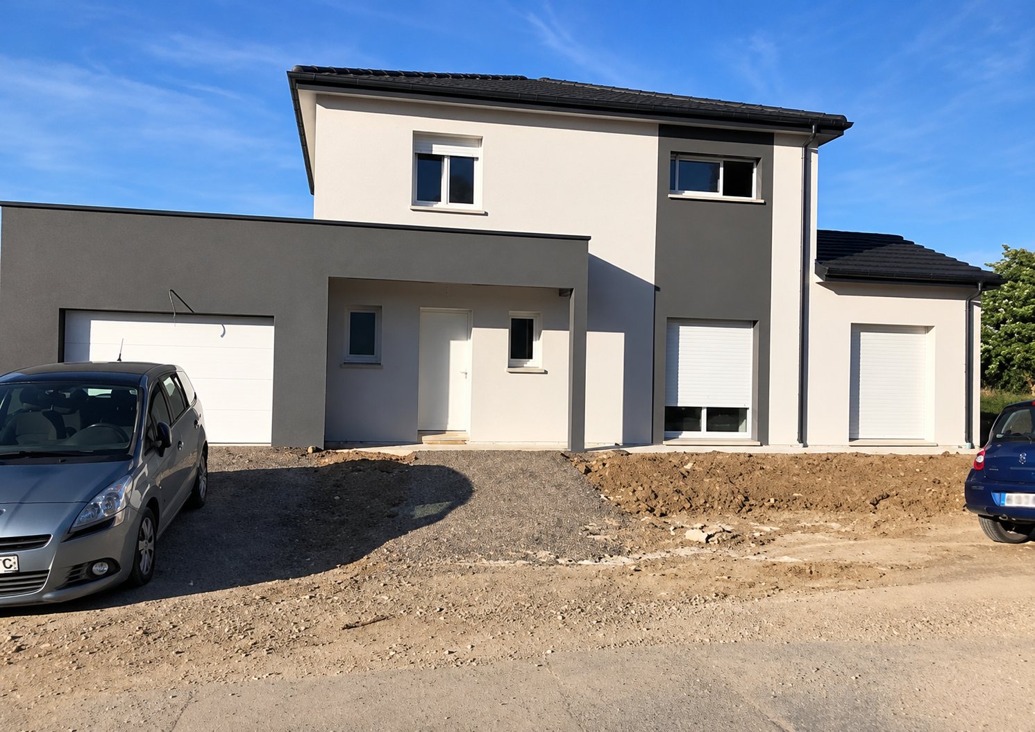 Maison individuelle récente à étage, aux façades blanches et grises, avec garage attenant et terrain en terre devant, où sont garées deux voitures.