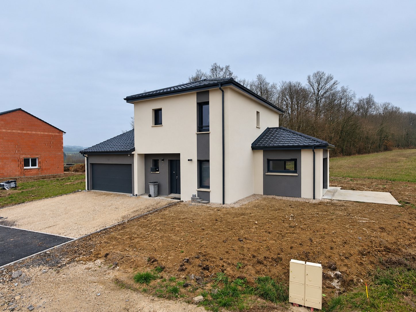 Maison individuelle neuve à étage, aux façades beige et grises et au toit en tuiles sombres, avec garage et terrain encore en terre.