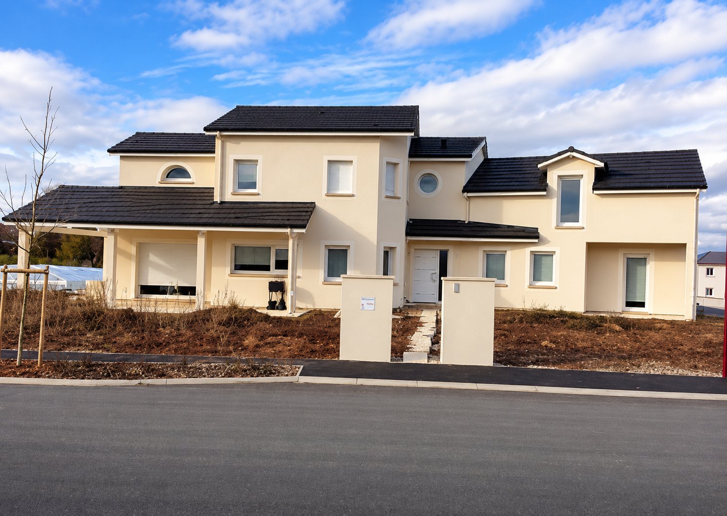 Grande maison individuelle récente à façade beige et toits en tuiles sombres, avec un terrain en cours d'aménagement devant une route.