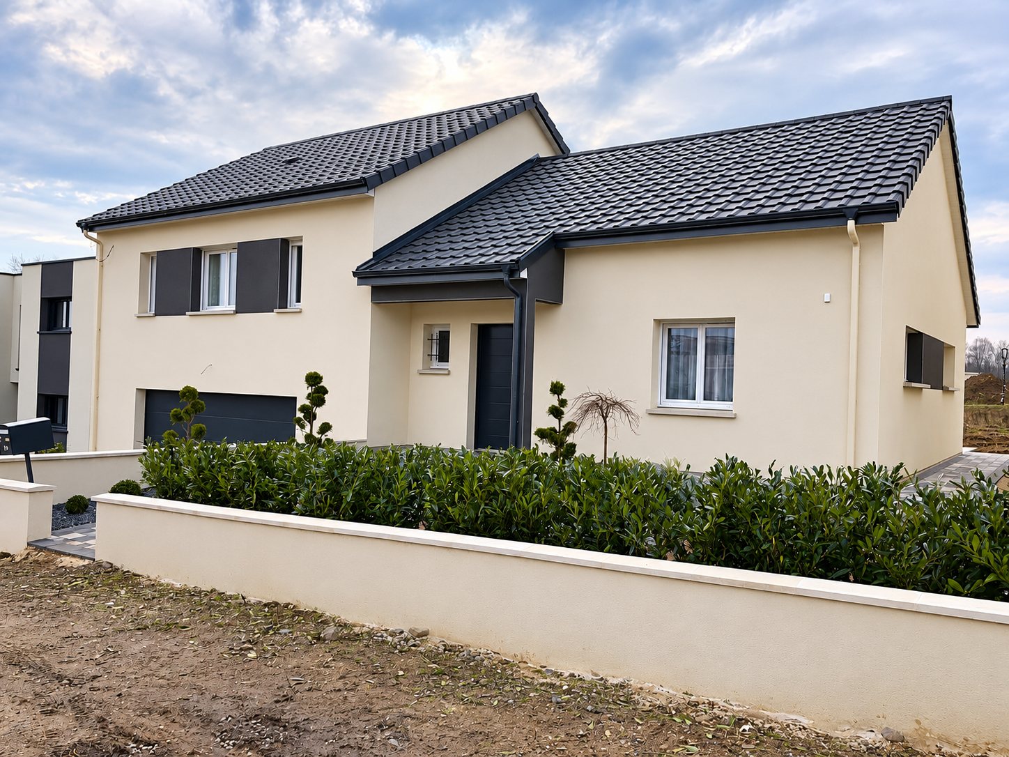 Maison individuelle récente aux façades beiges et à la toiture en tuiles gris foncé, entourée d'un muret blanc et d'une haie, sur un terrain encore en chantier.