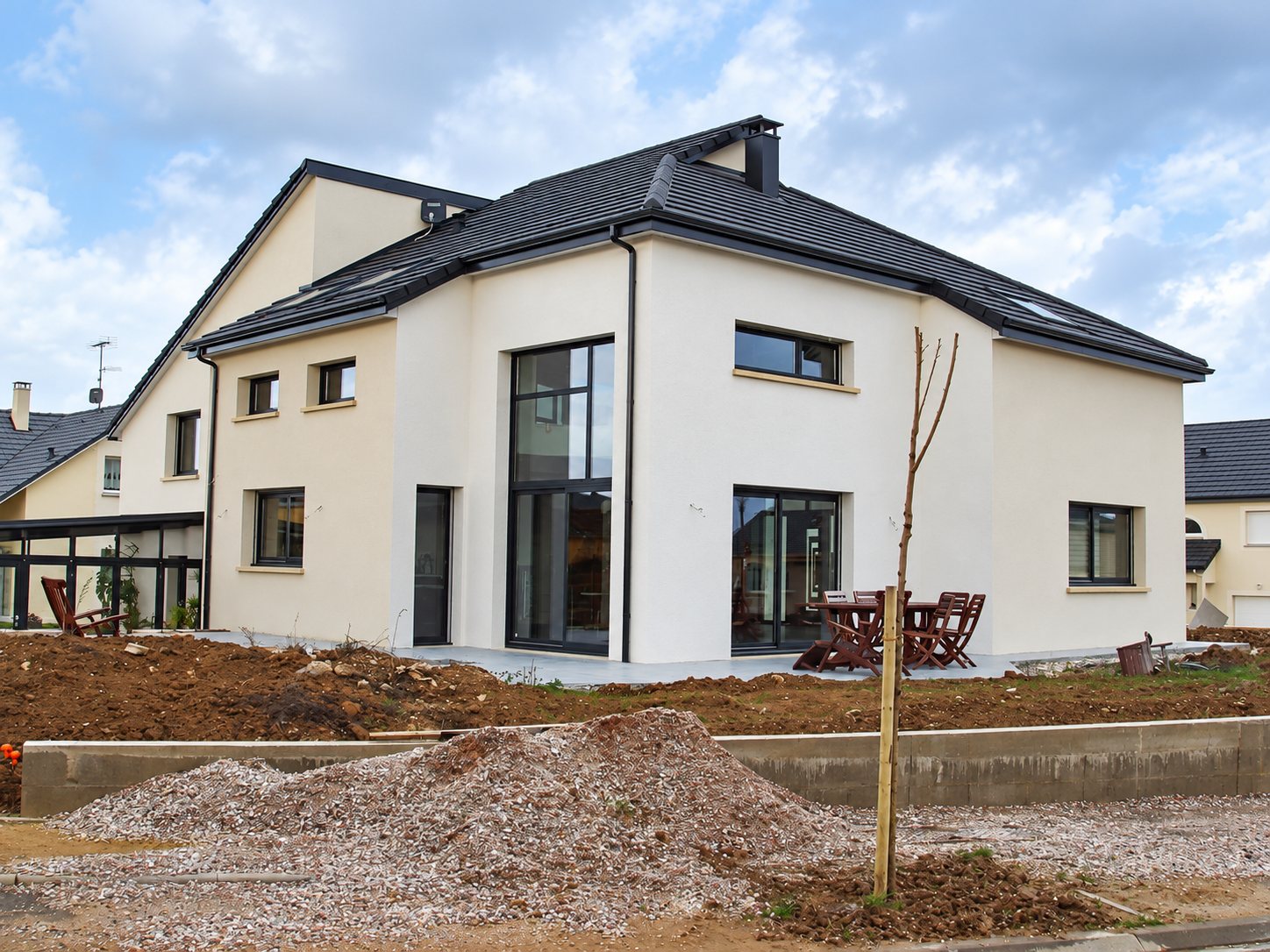 Maison individuelle récente aux murs blancs et au toit en tuiles sombres, entourée de terre fraîchement remuée et d'un tas de gravier sur un chantier de lotissement.