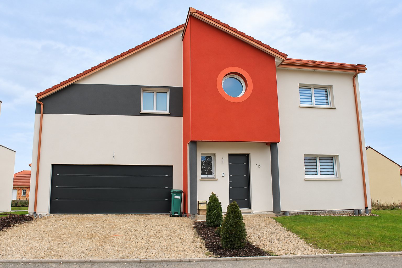 Façade d'une maison récente aux murs blancs, gris anthracite et rouge orangé, avec un hublot rond, une porte d'entrée sombre et un grand garage noir.