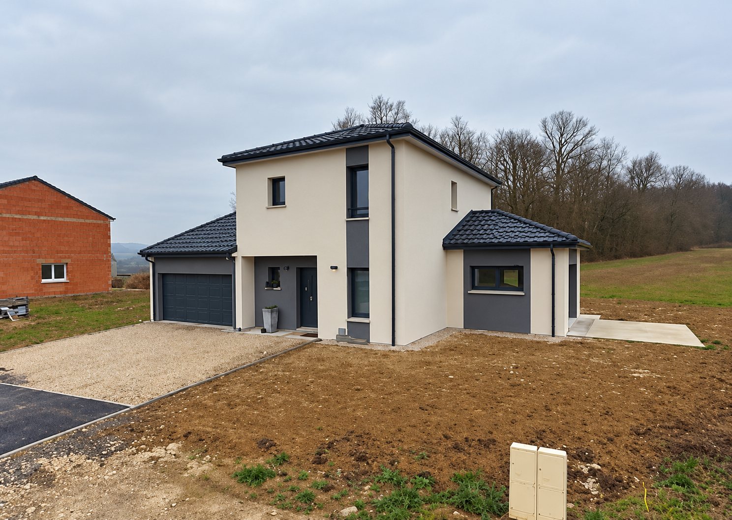 Maison individuelle neuve aux façades beige et gris avec toit en ardoise et garage, entourée d'un terrain en terre en cours d'aménagement.
