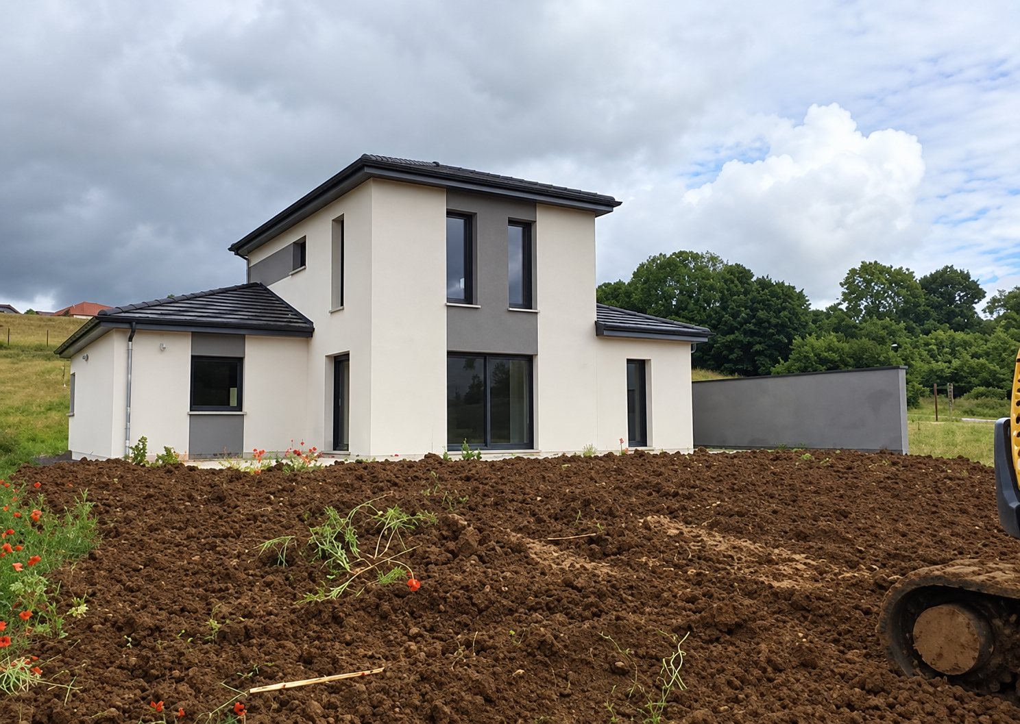 Une maison individuelle récente aux murs blancs et gris, avec un étage, entourée de terre fraîchement remuée sous un ciel nuageux.