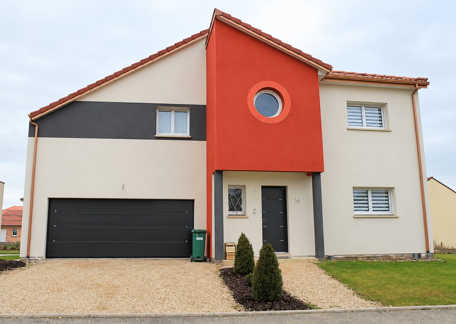 Maison individuelle récente à la façade crépie beige, grise et orange, avec un garage anthracite et une allée en gravier.