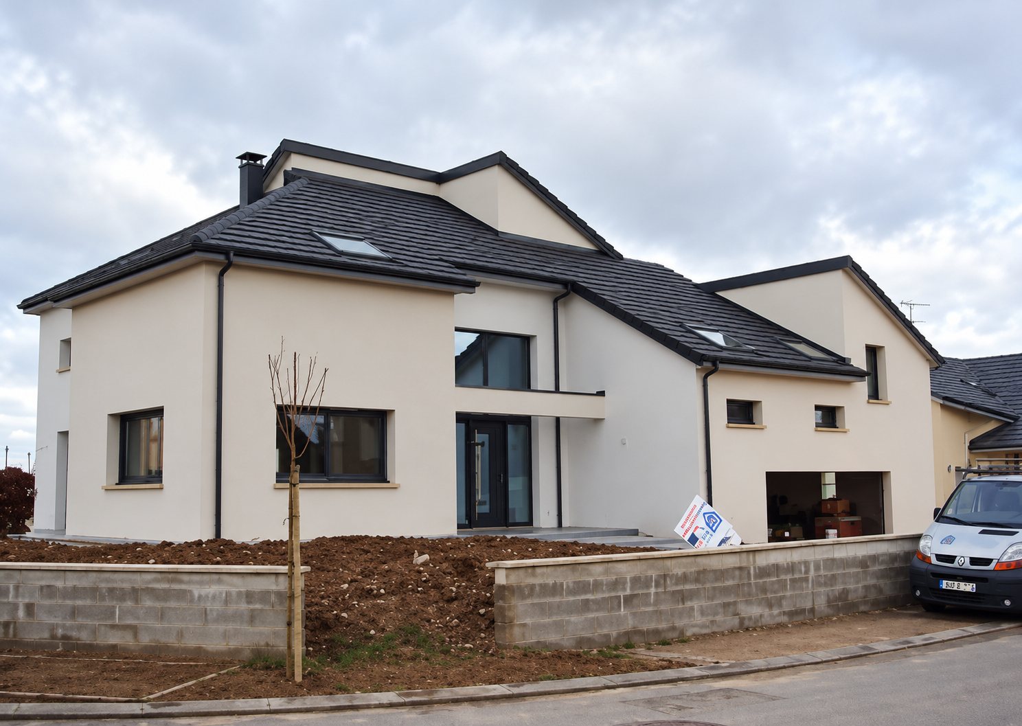 Une maison individuelle neuve aux murs enduits clairs et au toit sombre, entourée de terre fraîchement remuée, avec une camionnette blanche garée à droite.