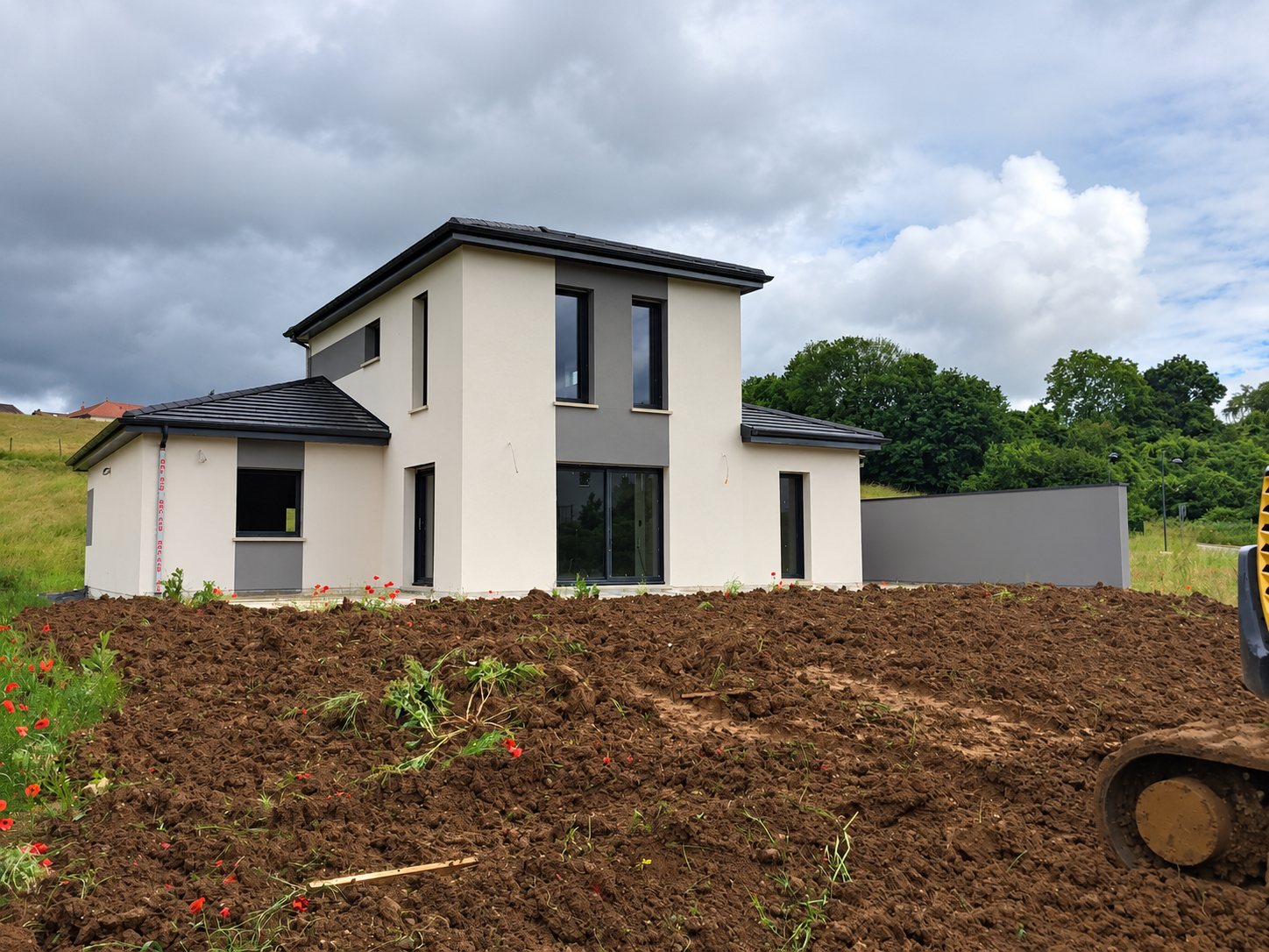 Une maison neuve à étage aux façades blanches et grises, entourée de terre fraîchement remuée, avec la chenille d'une pelleteuse au premier plan.