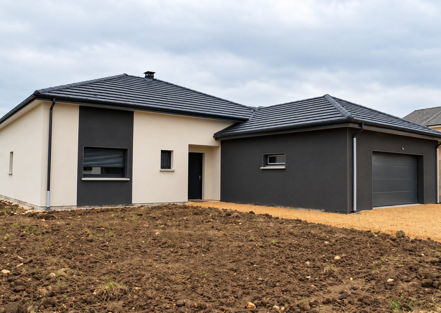 Maison individuelle récente de plain-pied aux murs blancs et gris anthracite, avec toit en tuiles sombres, garage et terrain en terre devant.