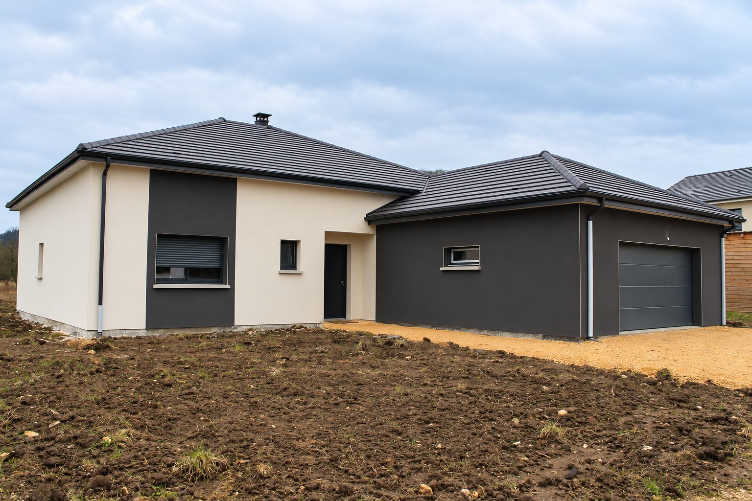 Maison individuelle récente de plain-pied aux façades beige et gris anthracite, avec garage attenant et terrain de terre fraîchement remuée au premier plan.
