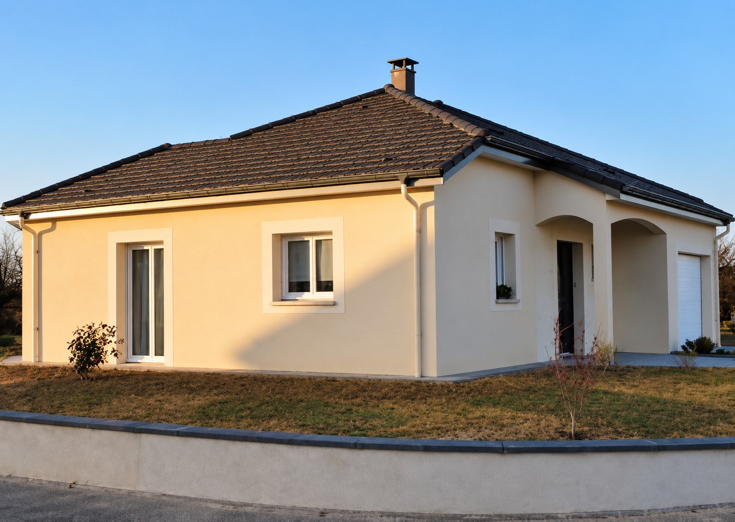 Maison récente de plain-pied à façade crépie beige, toit en tuiles sombres et garage, photographiée en fin de journée.