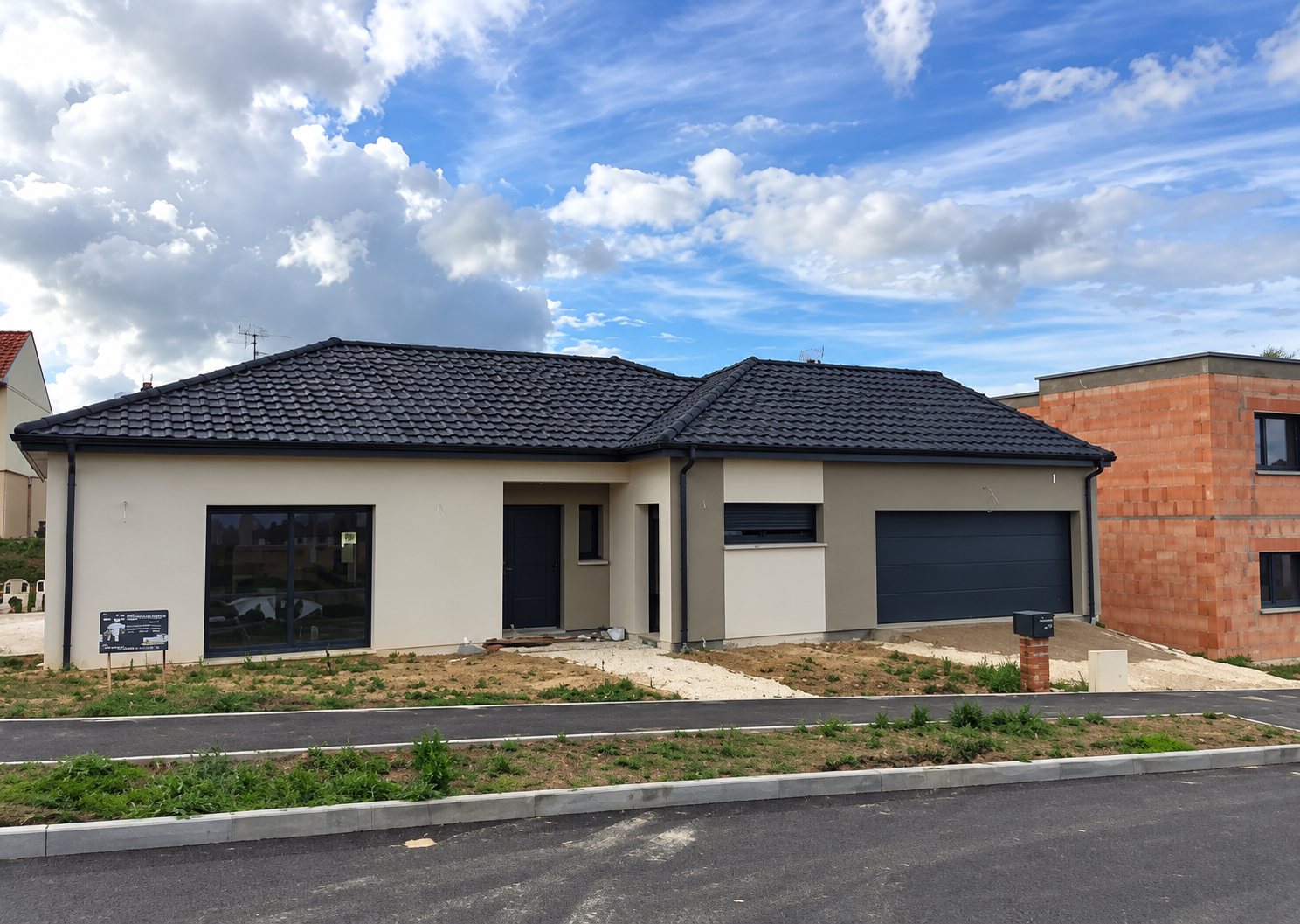 Une maison neuve de plain-pied aux murs clairs, au toit de tuiles anthracite et à la porte de garage grise, photographiée sous un ciel nuageux.