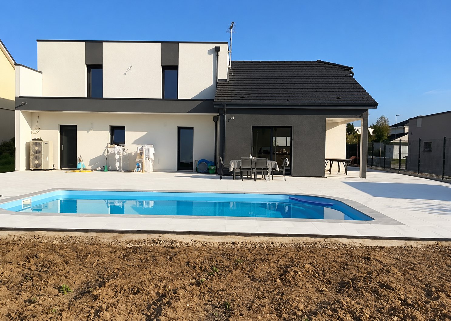 Une piscine bleue fraîchement installée entourée d'une terrasse claire devant une maison contemporaine à étage, avec un terrain de terre au premier plan.